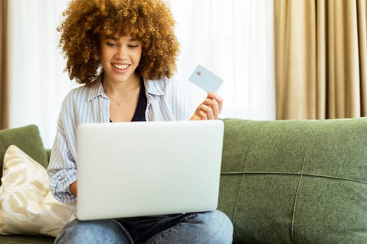 A young woman with curly hair happily shops online, holding a credit card, while seated on a comfortable couch in a stylish living room filled with warm sunlight.