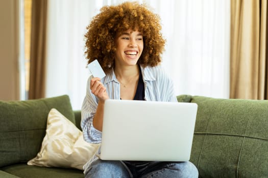A young woman with curly hair happily shops online, holding a credit card, while seated on a comfortable couch in a stylish living room filled with warm sunlight.