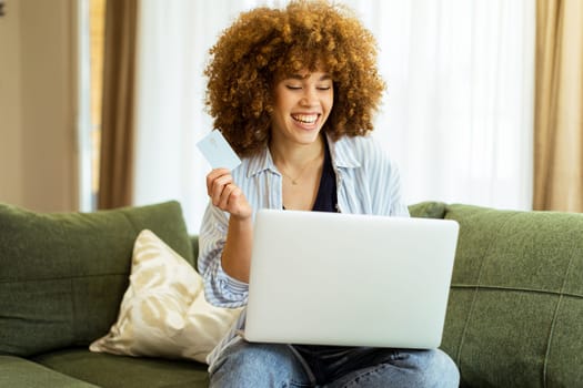 A young woman with curly hair happily shops online, holding a credit card, while seated on a comfortable couch in a stylish living room filled with warm sunlight.