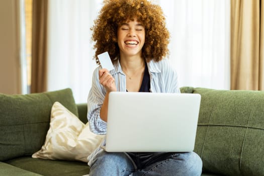 A young woman with curly hair happily shops online, holding a credit card, while seated on a comfortable couch in a stylish living room filled with warm sunlight.