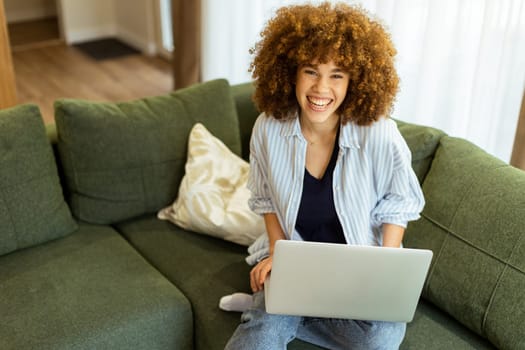 Sitting comfortably on a plush green sofa, a young woman types on her laptop, basking in the warm sunlight streaming through the large windows, radiating focus and tranquility.