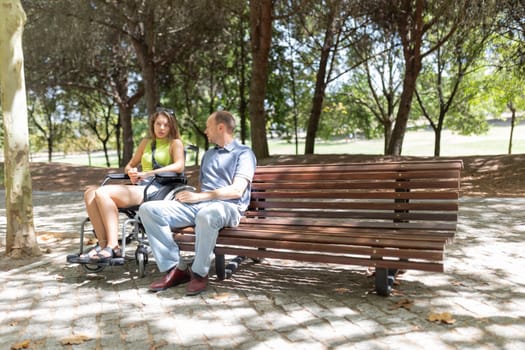 Young woman sitting in wheelchair and talking with a friend on a park bench