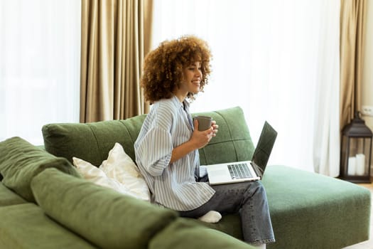 Sitting comfortably on a green sofa, a young woman savors a warm cup of coffee while working on her laptop in a bright, serene living room filled with natural light.