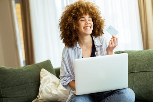 A young woman with curly hair happily shops online, holding a credit card, while seated on a comfortable couch in a stylish living room filled with warm sunlight.
