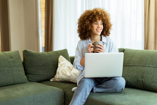 Sitting comfortably on a green sofa, a young woman savors a warm cup of coffee while working on her laptop in a bright, serene living room filled with natural light.