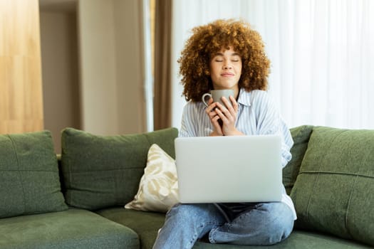 Sitting comfortably on a plush green couch, a young woman savors a steaming cup, embracing tranquility while working on her laptop in a sunlit home environment.