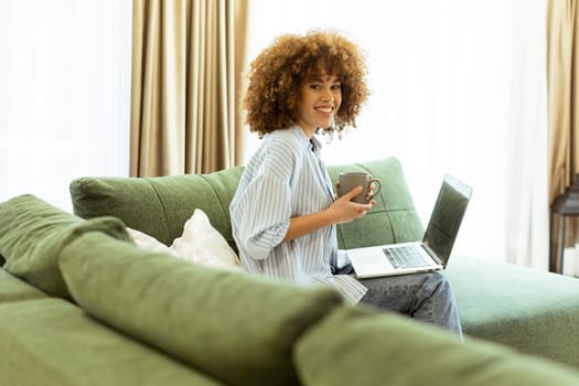 Sitting comfortably on a green sofa, a young woman savors a warm cup of coffee while working on her laptop in a bright, serene living room filled with natural light.