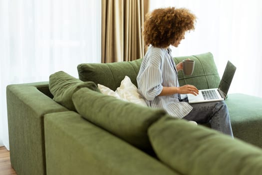 Sitting comfortably on a green sofa, a young woman savors a warm cup of coffee while working on her laptop in a bright, serene living room filled with natural light.