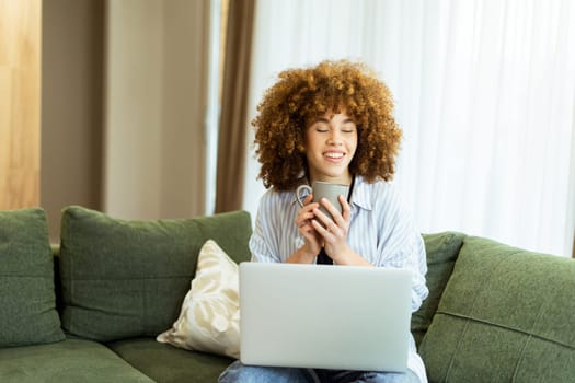 Sitting comfortably on a plush green couch, a young woman savors a steaming cup, embracing tranquility while working on her laptop in a sunlit home environment.