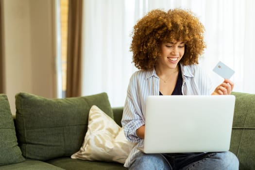A young woman with curly hair happily shops online, holding a credit card, while seated on a comfortable couch in a stylish living room filled with warm sunlight.
