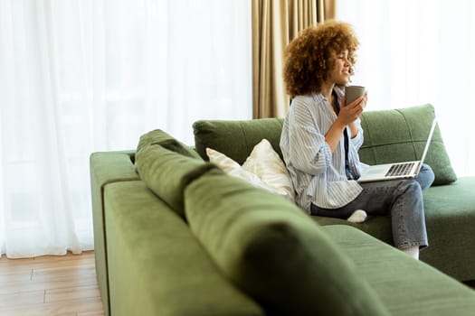 Sitting comfortably on a green sofa, a young woman savors a warm cup of coffee while working on her laptop in a bright, serene living room filled with natural light.