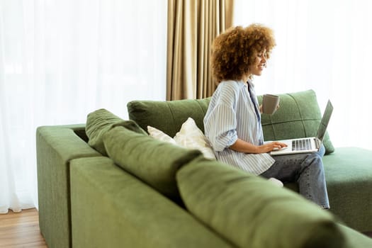 Sitting comfortably on a green sofa, a young woman savors a warm cup of coffee while working on her laptop in a bright, serene living room filled with natural light.
