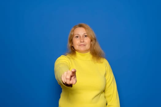Caucasian woman in her 40s wearing a yellow sweater pointing at the camera while smiling in a positive attitude, isolated over blue studio background