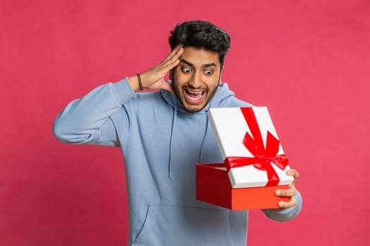 Excited Indian man receiving a festive gift with a red bow opening the box with joy celebrating a surprise. Happy Arabian guy enjoying dream present smiling with excitement isolated on red background