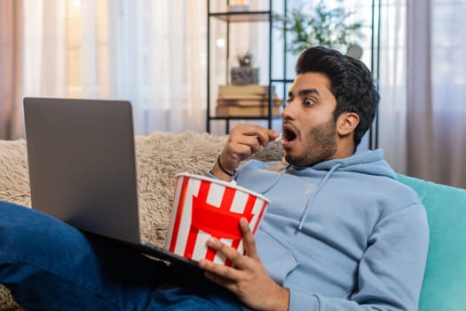 Indian young man eating popcorn and watching movie on laptop, enjoying relaxed time. Arabian guy sits on sofa in living room smiling while having snacks and staying entertained with favorite film.