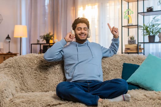 Indian young man sitting on sofa couch listening to music through headphones dances to rhythm relaxes and unwinds. Hispanic guy enjoys beats moving with energy and finding joy in favorite tracks.