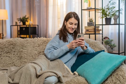 Happy young woman in casual clothes text messaging on smartphone. Caucasian girl covered with blanket lying on couch and using mobile phone in living room at home. Smiling lady relaxing in apartment.