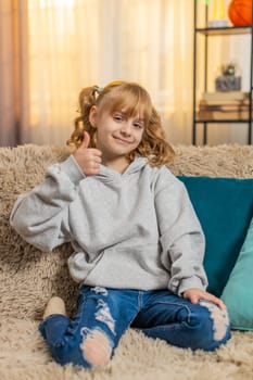 Teenager girl showing thumbs up like with a cheerful expression at home, reflecting happiness and satisfaction. Caucasian child kid enjoying success and expressing approval while sitting on a sofa.
