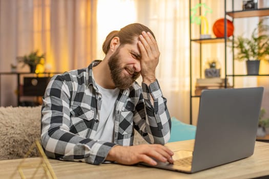 Man concentrates at home table, typing on laptop until stumbling upon a big glitch error virus that sparks immediate annoyance. Guy slaps forehead, quietly grumbling at unexpected technical issue fail
