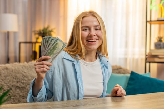 Young woman sits at home counting money dollar cash in hand, happy laughter from sudden lottery jackpot victory, salary, earnings. Caucasian girl waves banknotes celebrating financial surprise. Sunset