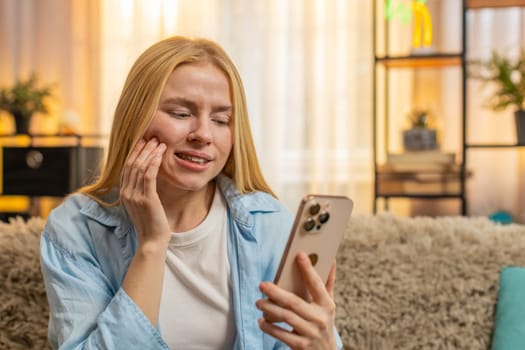 Young woman holding cheek at home, showing tension and urgent need for dental care. Girl scrolling through smartphone with painful expression, toothache and anxiety, searching for nearest dentistry