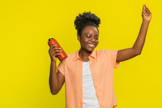 African American woman holds red portable speaker, moving energetically to the rhythm of music with ease. Black girl isolated on yellow background showing joy relaxation while enjoying favorite tunes