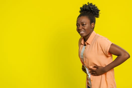 African American woman holding stomach with both hands, grimacing in pain discomfort. Black girl isolated on yellow background abdominal distress from overeating, gastritis indigestion, menstrual pain