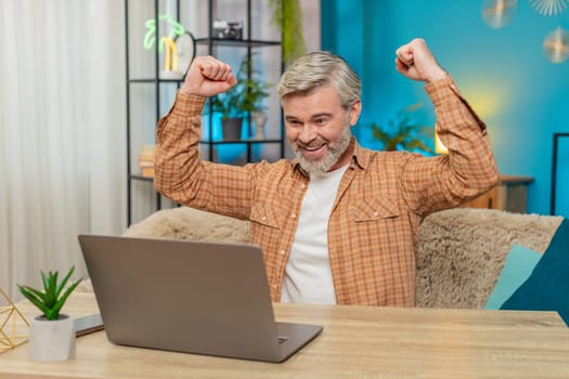 Happy Caucasian mature man using laptop achieve an important email message good news. Satisfied middle-aged guy sitting at home table joyfully exclaims, raising his hands in victory after winning game