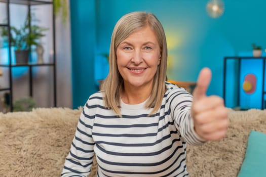 Like sign. Caucasian mature woman gives thumbs up sitting on home sofa, smiling and showing approval support. Grandmother expresses positivity and encouragement, positive feedback, good great news