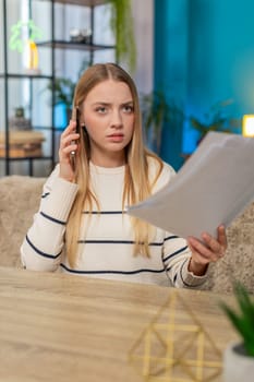 Caucasian woman tries to solve urgent issue at home at table, talking on phone, holding papers graphs, sounding distressed. Girl feels worried frustrated searching answers seeking help feeling anxious