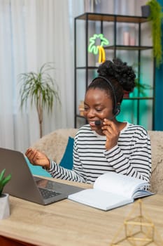 African American Black woman call center operator sitting on sofa at home with headset focused on work writing notes in notebook. Calm attentive girl working remotely in living room apartment setting.