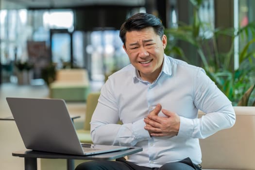 Stressed Chinese mature businessman using laptop and suffers from tension holding chest with heart pain attack risk of stroke symptom while working in modern office lobby. Businessperson in shirt.