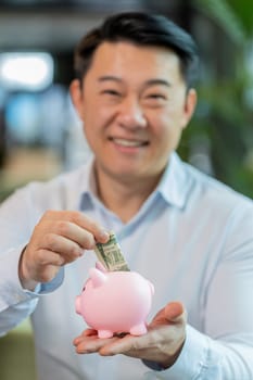 Happy mature businessman entrepreneur save money using pink piggybank putting dollar cast inside. Success, economy, investment, deposit. Guy manager in shirt sitting with laptop in modern office lobby