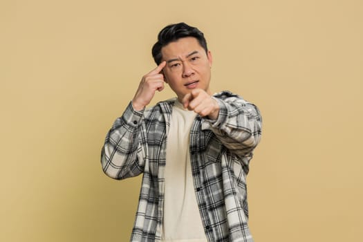 You are crazy, out of mind. Confused Asian middle-aged man pointing at camera and showing stupid gesture, blaming asking some idiot for insane plan, bullying. Mature Chinese guy on beige background