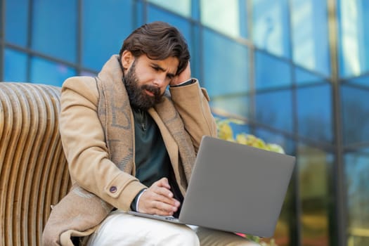 Sad Arabian man tourist using laptop computer typing browsing, loses becoming surprised sudden lottery results, bad news, fortune loss, fail. Angry guy sitting on bench downtown. Town lifestyles.