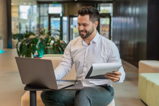 Caucasian young businessman using laptop and working with documents, analyzing information, reading papers prepare financial report. Male entrepreneur in formal analyzing graph in modern office lobby.
