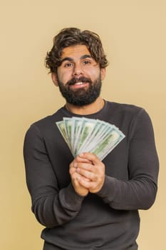 Rich pleased boss handsome young man waving money dollar cash banknotes bills like a fan, success business career, lottery winner, big income, wealth. Adult Indian guy isolated on beige background.