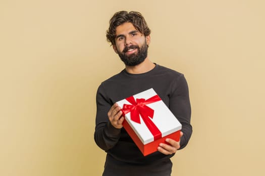 Positive smiling young Indian man presenting birthday gift box stretches out hands, offer wrapped present career bonus, celebrating party, sale. Bearded Hindu guy isolated on beige studio background.