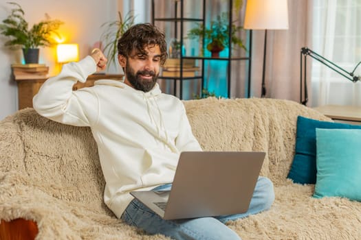 Excited young Indian man guy using laptop computer, receive mail good news message, wow shocked by sudden victory celebrate lottery jackpot win purchases online shopping at home on sofa. Lifestyles.
