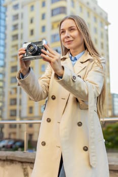 Happy Caucasian middle-aged woman tourist traveling on weekends exploring city making photo pictures on retro vintage camera outdoors. Girl in long coat on urban street during vacation. Lifestyles.