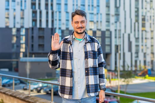 Cheerful mature man tourist smiling friendly at camera, waving hands gesturing invitation hello, hi, greeting, goodbye, welcoming with hospitable expression outdoors. Happy guy on downtown city street