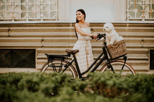 Pretty young woman with white bichon frise dog in the basket of electric bike