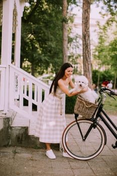 Pretty young woman putting white bichon frise dog in the basket of electric bike