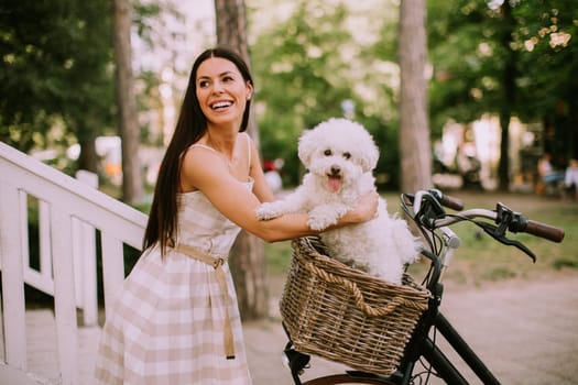 Pretty young woman putting white bichon frise dog in the basket of electric bike