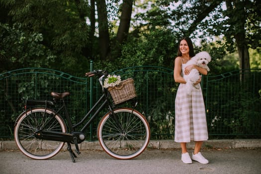 Pretty young woman putting white bichon frise dog in the basket of electric bike