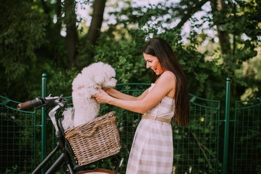 Pretty young woman putting white bichon frise dog in the basket of electric bike