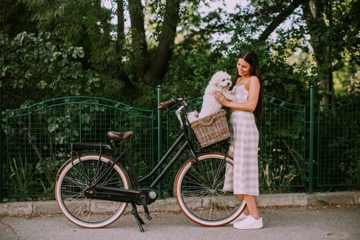 Pretty young woman putting white bichon frise dog in the basket of electric bike