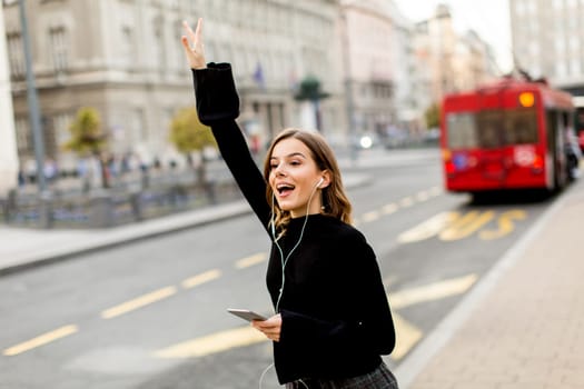 Smiling brunette woman using mobile while standing on street and  waiting for a bus or taxi