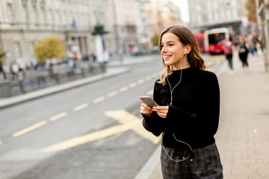 Smiling brunette woman using mobile while standing on street and  waiting for a bus or taxi