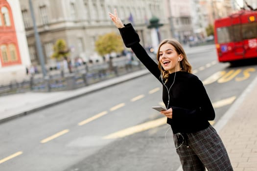 Smiling brunette woman using mobile while standing on street and  waiting for a bus or taxi